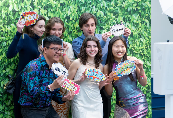 students pose in photobooth with green background holding various signs