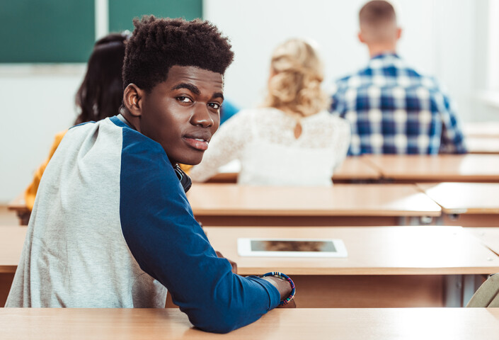 Smiling Black male student seated at a desk in a classroom looks over his right shoulder