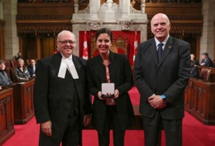 Melanie Adrian holding her medal from the Canadian Senate, with Speaker of the Senate George Furey to her left and Senator Vernon White to her right.