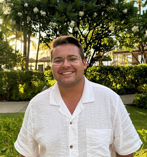 Picture of a young man with glasses, brown hair, and a white buttoned down shirt in front of a sunny outdoor background