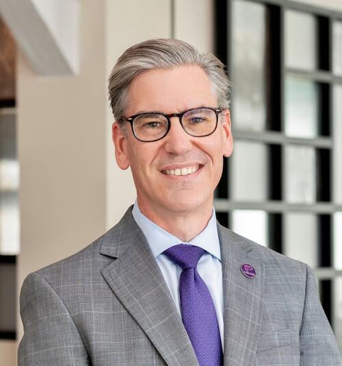 Headshot of man in gray suit, purple tie, wearing eyeglasses and has short grey hair