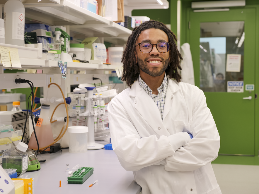 Student smiling with crossed arms, wearing white lab coat, standing next to lab bench