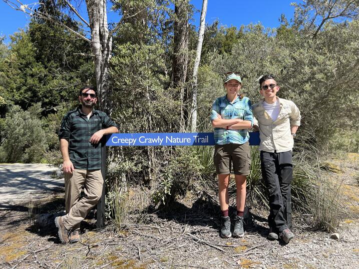 Three smiling people in nature next to a sign that says "Creepy Crawly Nature Trail"