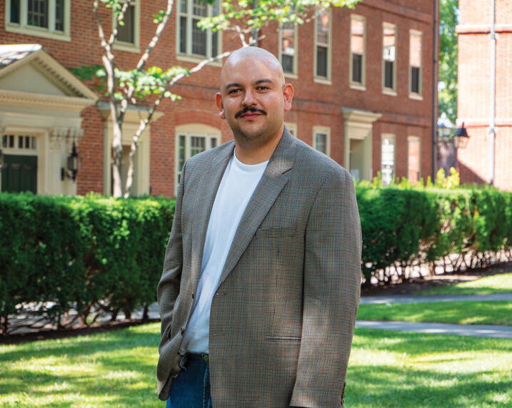 Headshot of Marco Avina wearing gray suit jacket, white shirt, standing in front of brick building