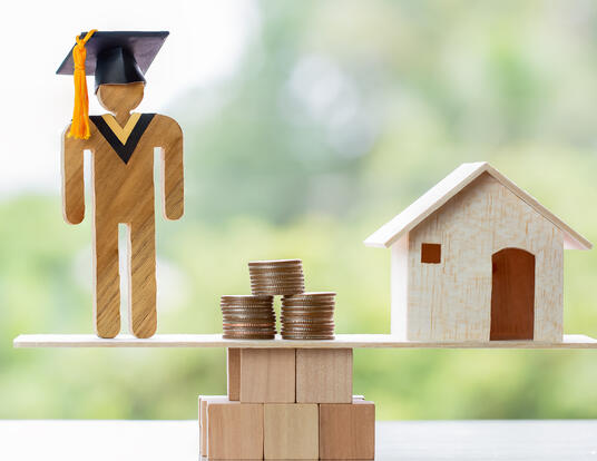 wooden silhoutte of graduate next to stacks of coins and a house