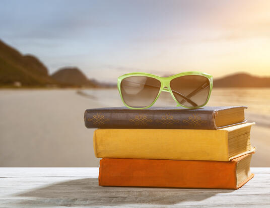 pile of old books on a table, with sunglasses on top and a beach in the background