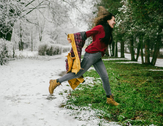 girl jumping in the snow from green grass wearing boots and winter coat