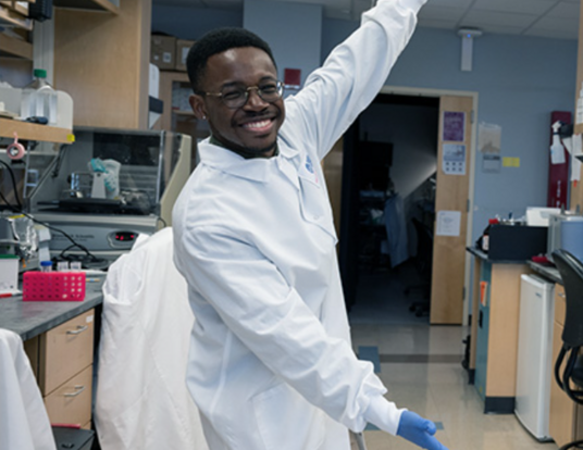 Olúmídé Fagboyegun smiling and gesturing toward his lab equipment