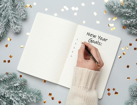 Woman writing in notebook at light grey table with Christmas decor, top view. New Year Goals