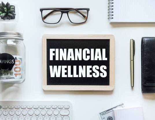 Financial Wellness written on a screen surrounded by glasses, a jar with money, a pen, notebook, and laptop keyboard.