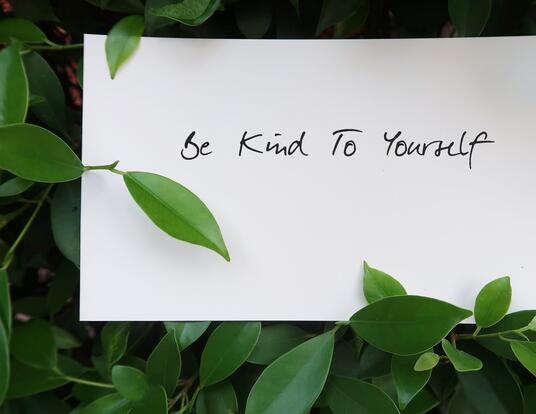paper that reads, "Be Kind To Yourself" with green leaves around it