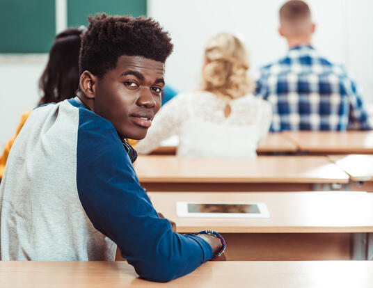 Smiling Black male student seated at a desk in a classroom looks over his right shoulder 
