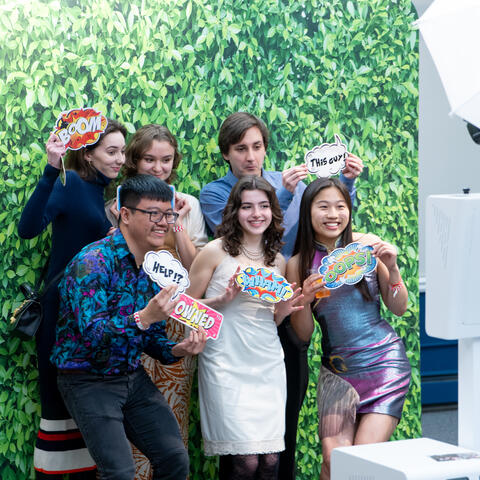 students pose in photobooth with green background holding various signs