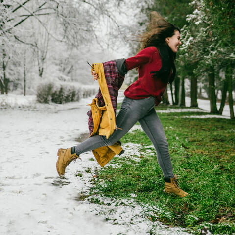 girl jumping in the snow from green grass wearing boots and winter coat