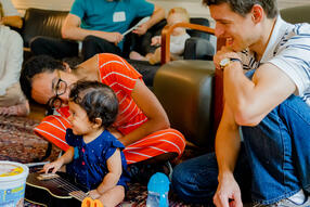 Smiling child holding guitar and two smiling adults all sitting on the floor