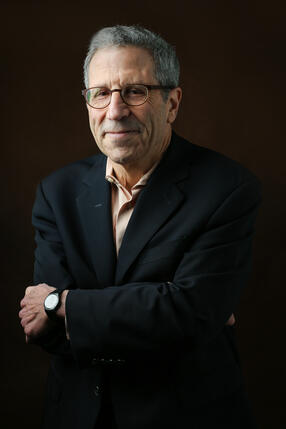 male with black suite, tie, glasses, arms crossed in front of him
