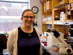 Maddy Mcfarland, wearing a blue shirt and tan sweater, smiles in front of lab equipment