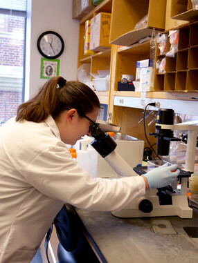 Maddy Mcfarland, wearing a white lab coat, looks into a microscope