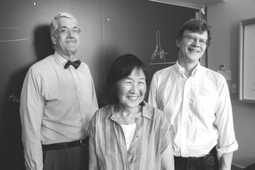 John Doyle (in a button down and bow tie), Evelyn Hu (smiling and wearing a button down shirt), and Mikhail Lukin (also smiling and wearing a button down shirt), stand in front of a blackboard with a roughly drawn chalk graph