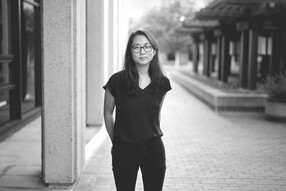 Di Wei, wearing a t-shirt and glasses, stands outdoors with her hands behind her back