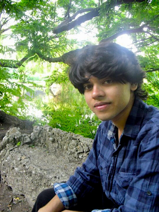 Amar Sarkar profile shot wearing a black and blue plaid shirt; green treats and a rock in the background