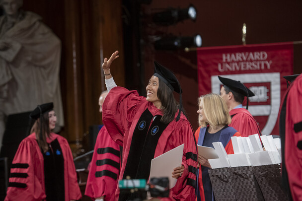 Smiling woman stands in academic regalia and waves from the stage