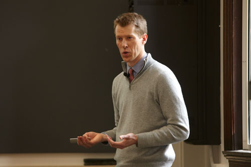 man in gray sweater, blue shirt, and red tie speaking at front of classroom