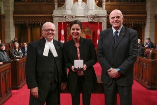 Melanie Adrian holding her medal from the Canadian Senate, with Speaker of the Senate George Furey to her left and Senator Vernon White to her right.